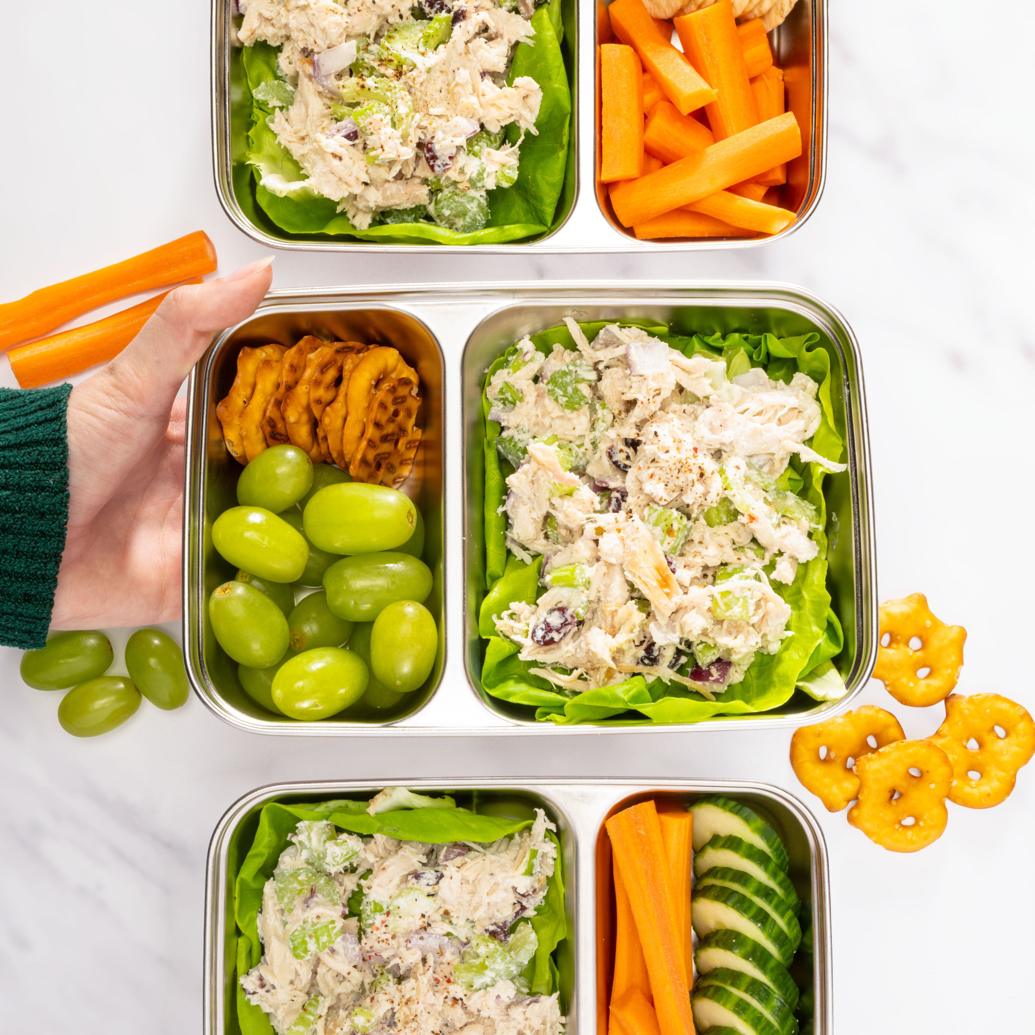 two compartments of a bento box with chicken salad, grapes, carrots, and crackers on a marble surface.